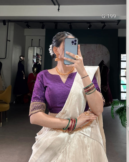 Woman taking a mirror selfie wearing a purple blouse and white saree with jewelry.