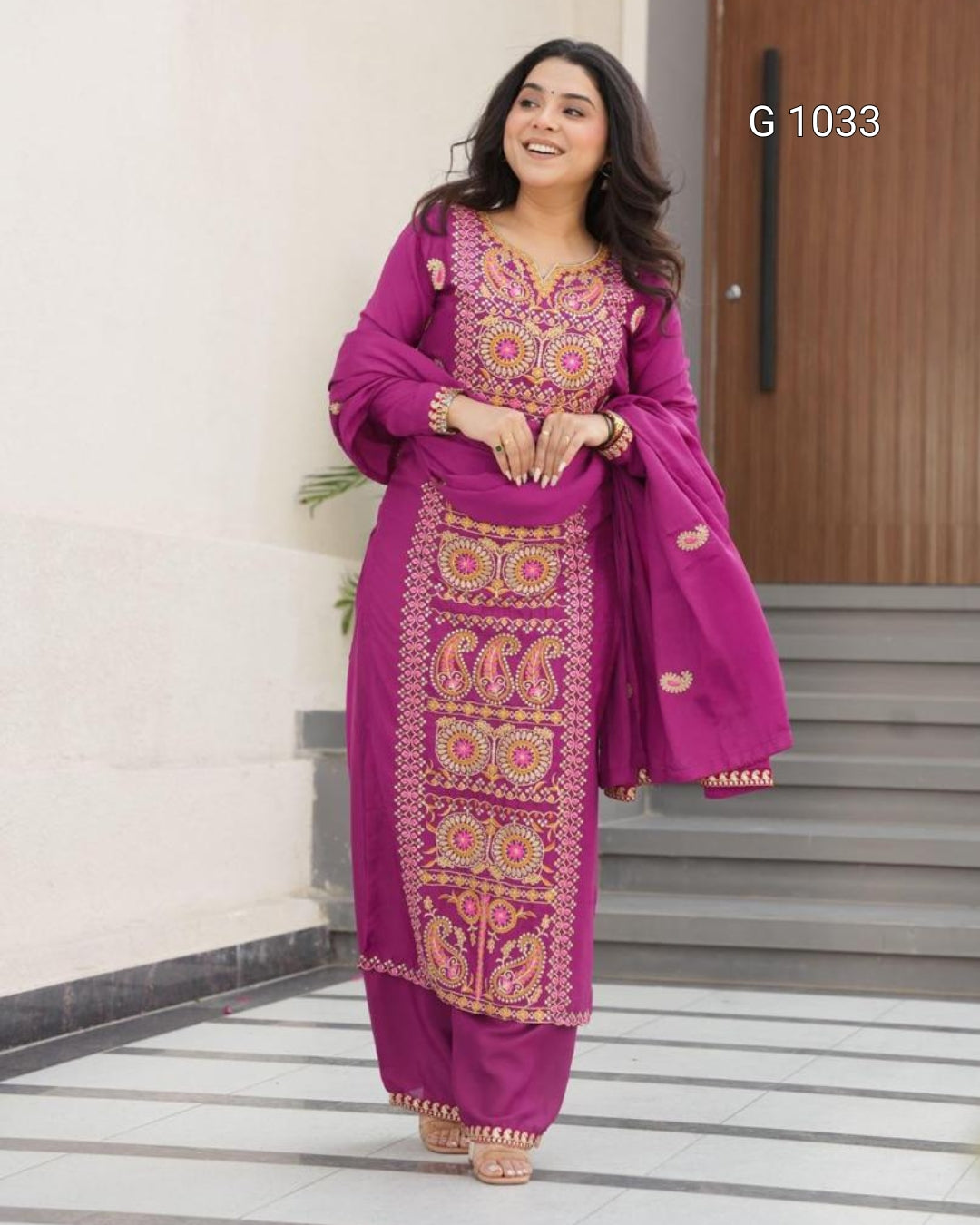 A woman modeling a magenta purple ethnic salwar kameez suit with intricate gold and green embroidery, standing on outdoor concrete steps.