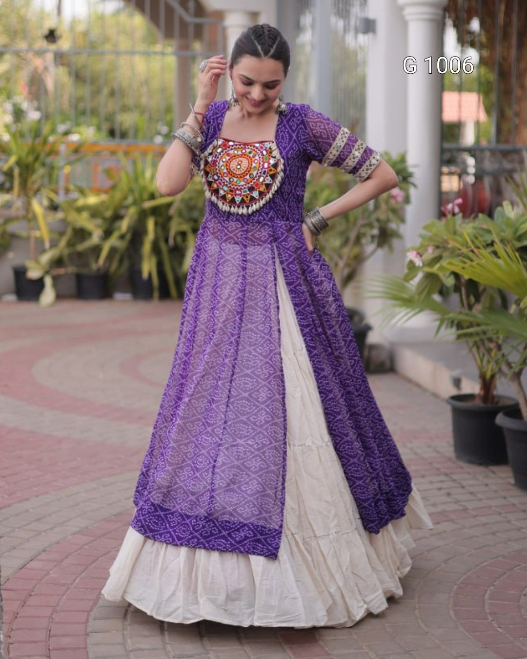 A woman wearing a traditional Indian outfit with a purple embroidered kurta and a white flared skirt, standing in an outdoor courtyard with potted plants.