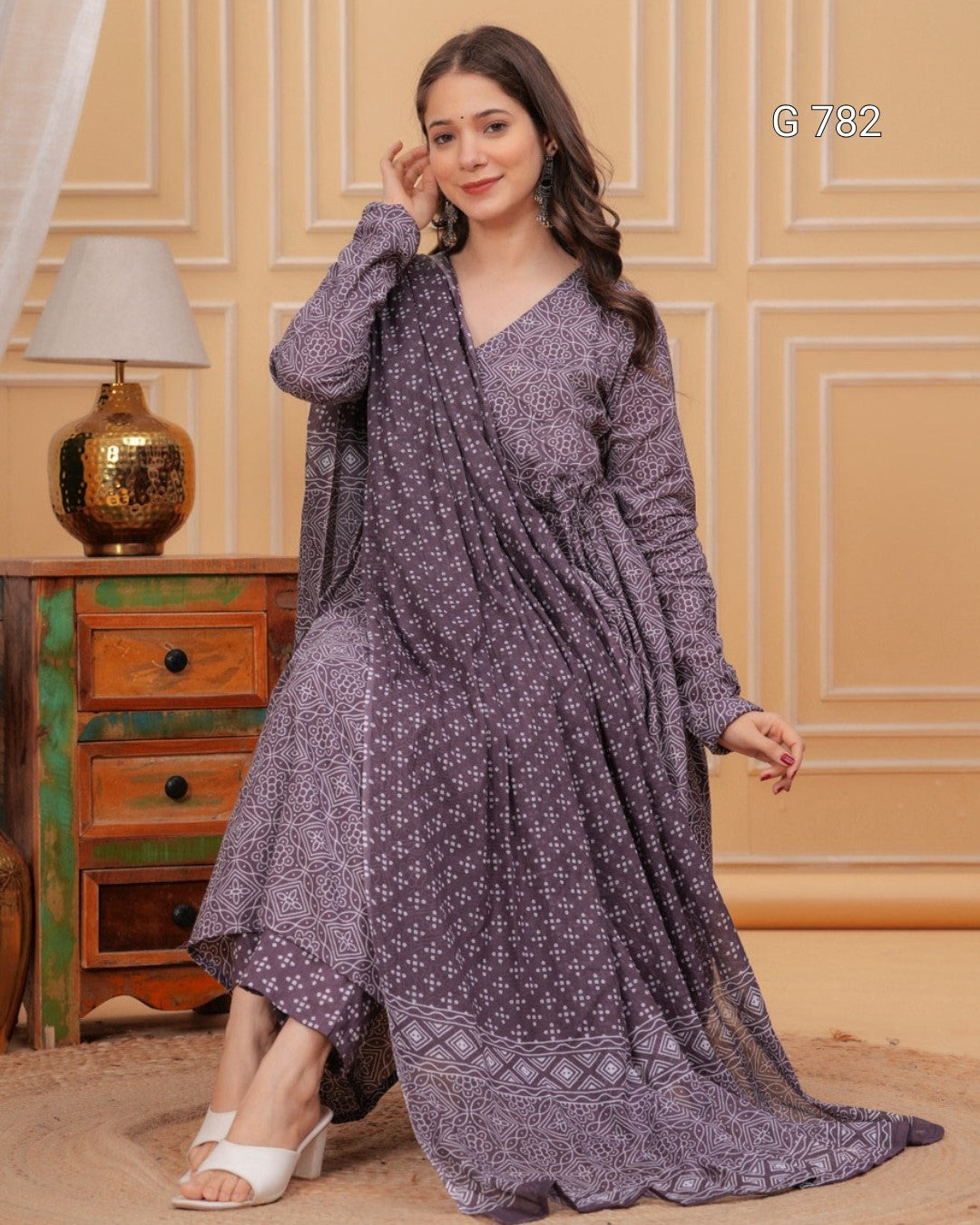Woman wearing a long, patterned dress sitting on a wooden floor with a decorative wall in the background.