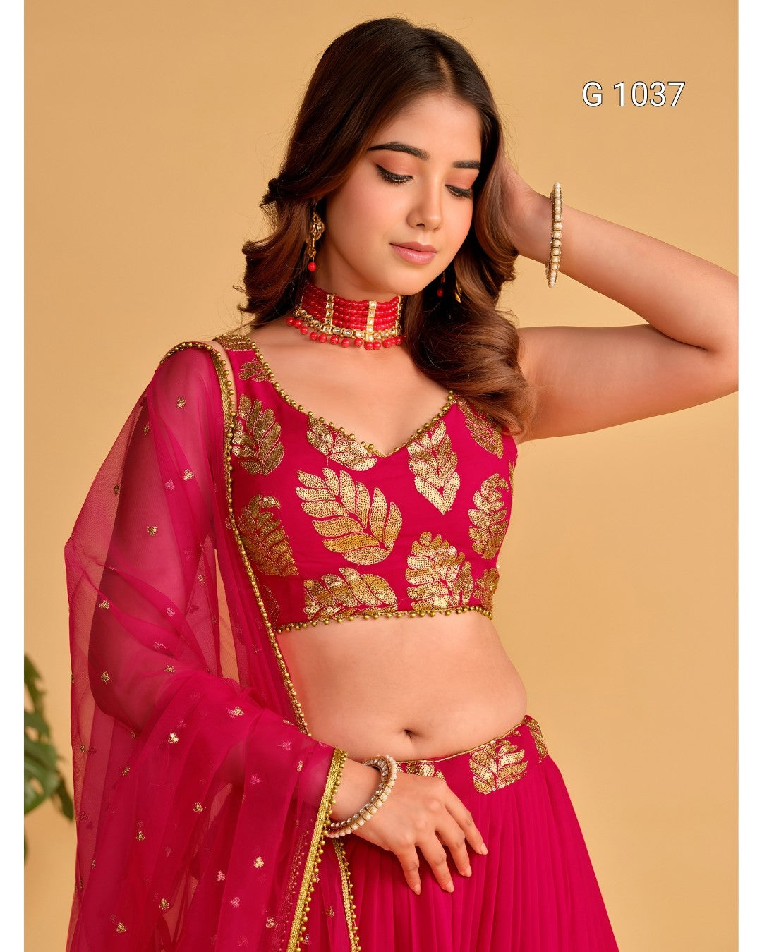 A woman modeling a bright pink traditional outfit with gold embroidered leaf patterns on the blouse and matching sheer dupatta, set against a solid beige background.