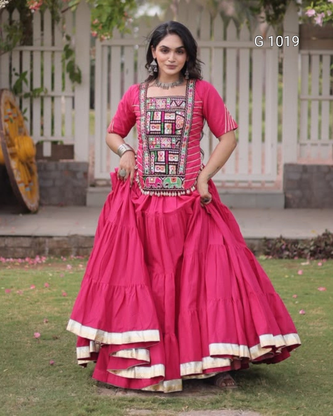 A woman modeling a bright pink traditional Indian dress with a heavily embroidered bodice and a voluminous tiered skirt, standing outdoors in front of a white fence and green lawn.