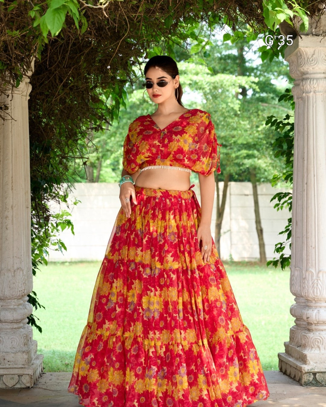 Woman in a red and yellow floral outfit standing under a stone archway with greenery in the background.