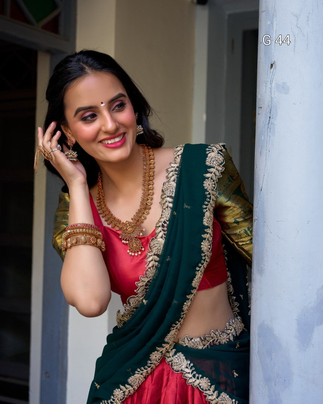 Woman in traditional green and red saree with jewelry, standing indoors.