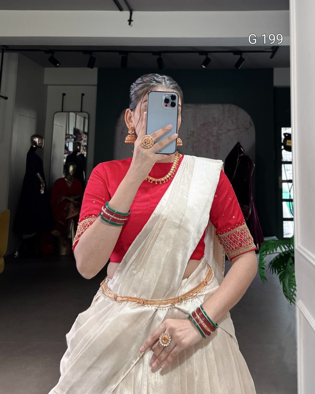 Person wearing a red blouse and white saree with jewelry, taking a mirror selfie.