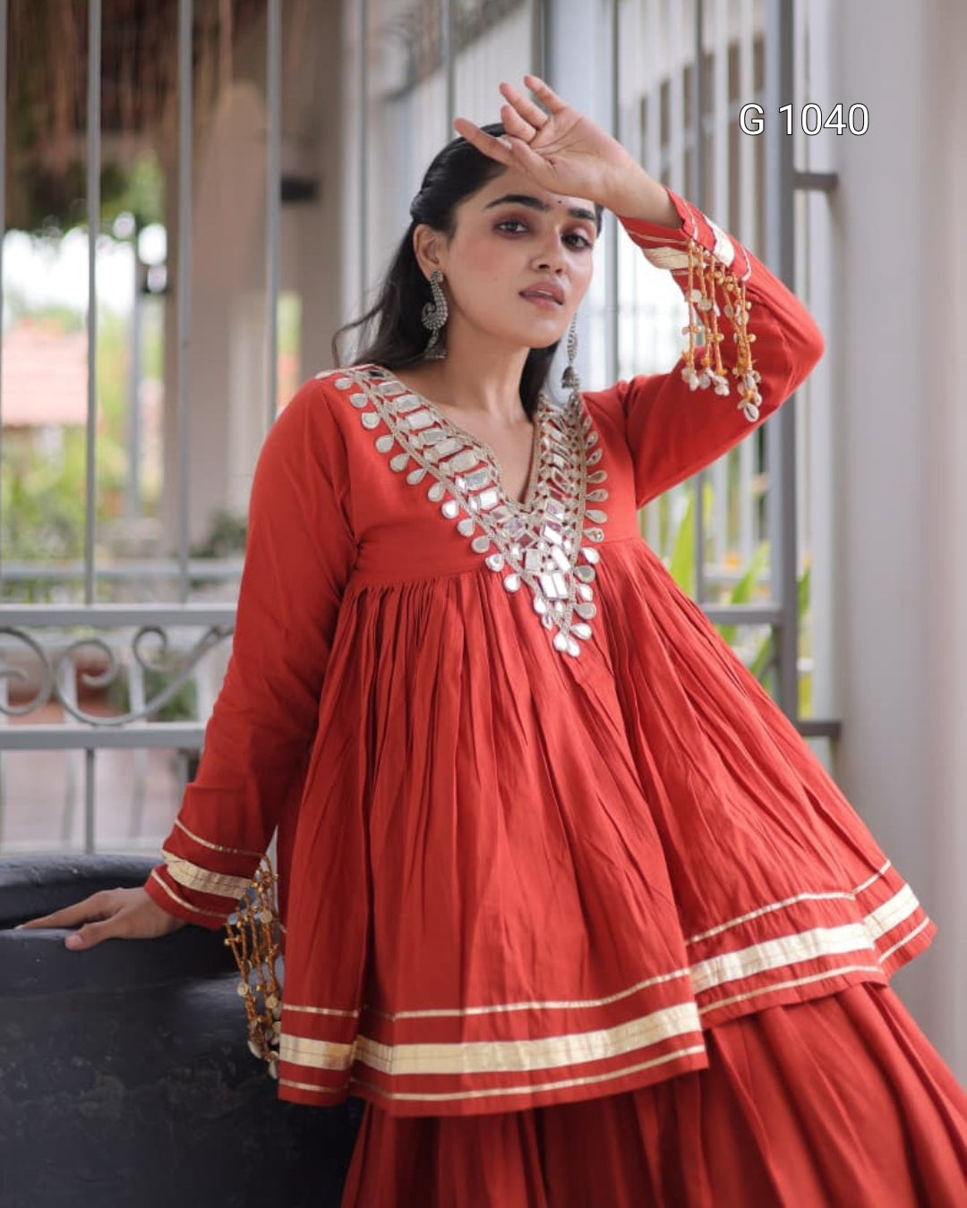 A woman models a red traditional Anarkali-style dress with gold and silver mirror work embroidery on the neckline and sleeve cuffs, set against a porch background.