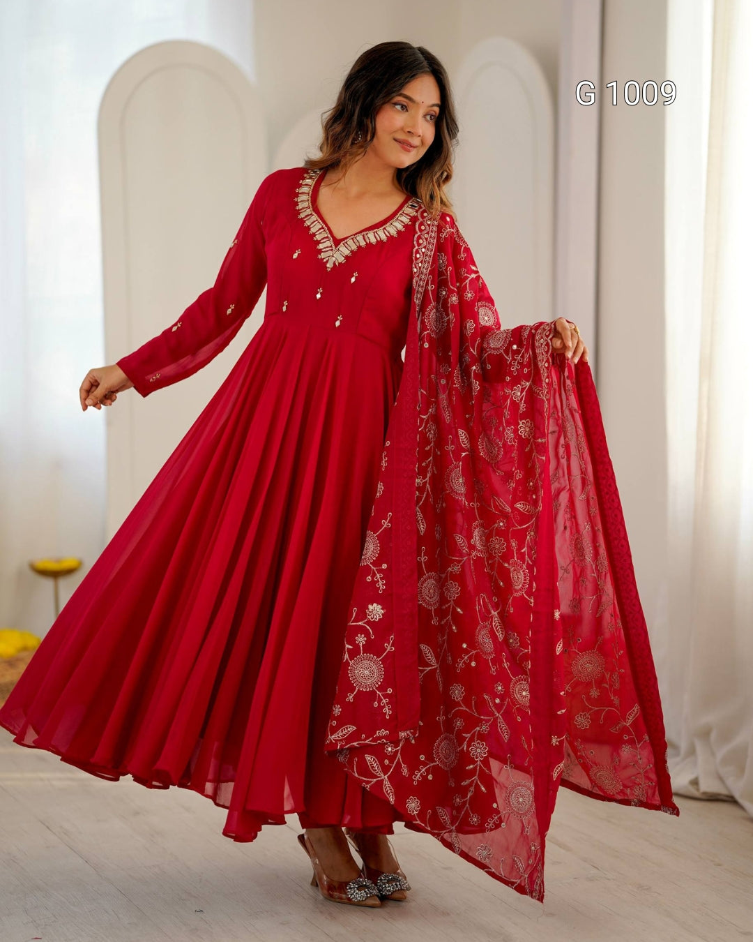 A woman modeling a long red anarkali-style dress with silver floral embroidery and a matching sheer red dupatta draped over one arm, shown in a bright indoor studio setting.