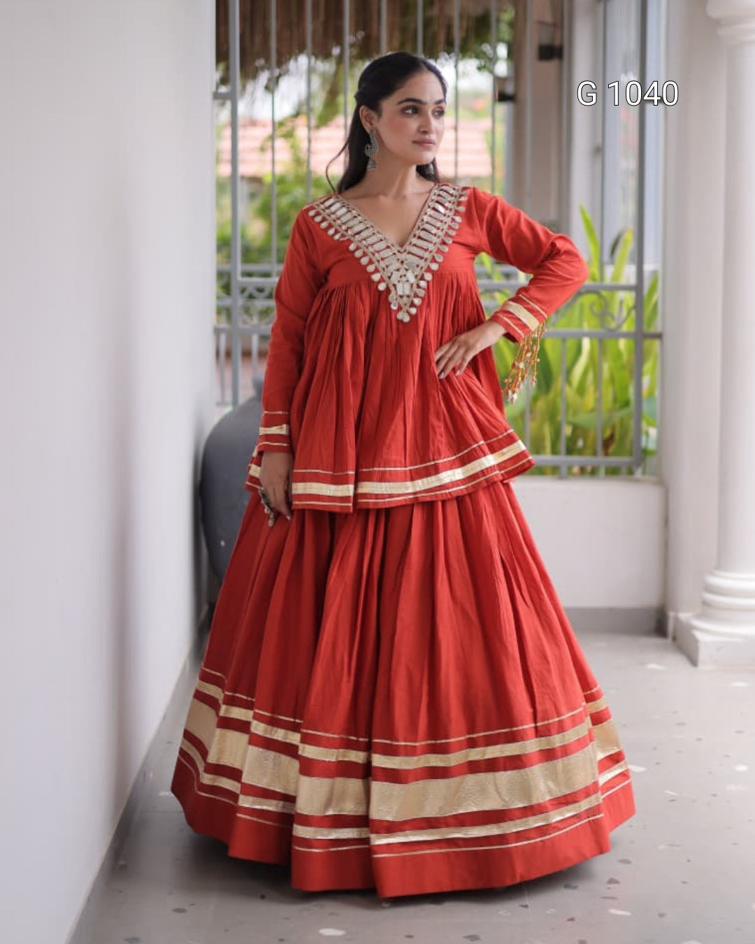 A woman models a traditional Indian outfit consisting of a red long kurta and a matching flared skirt with beige and white striped borders, set against a white-walled corridor with a window in the background.