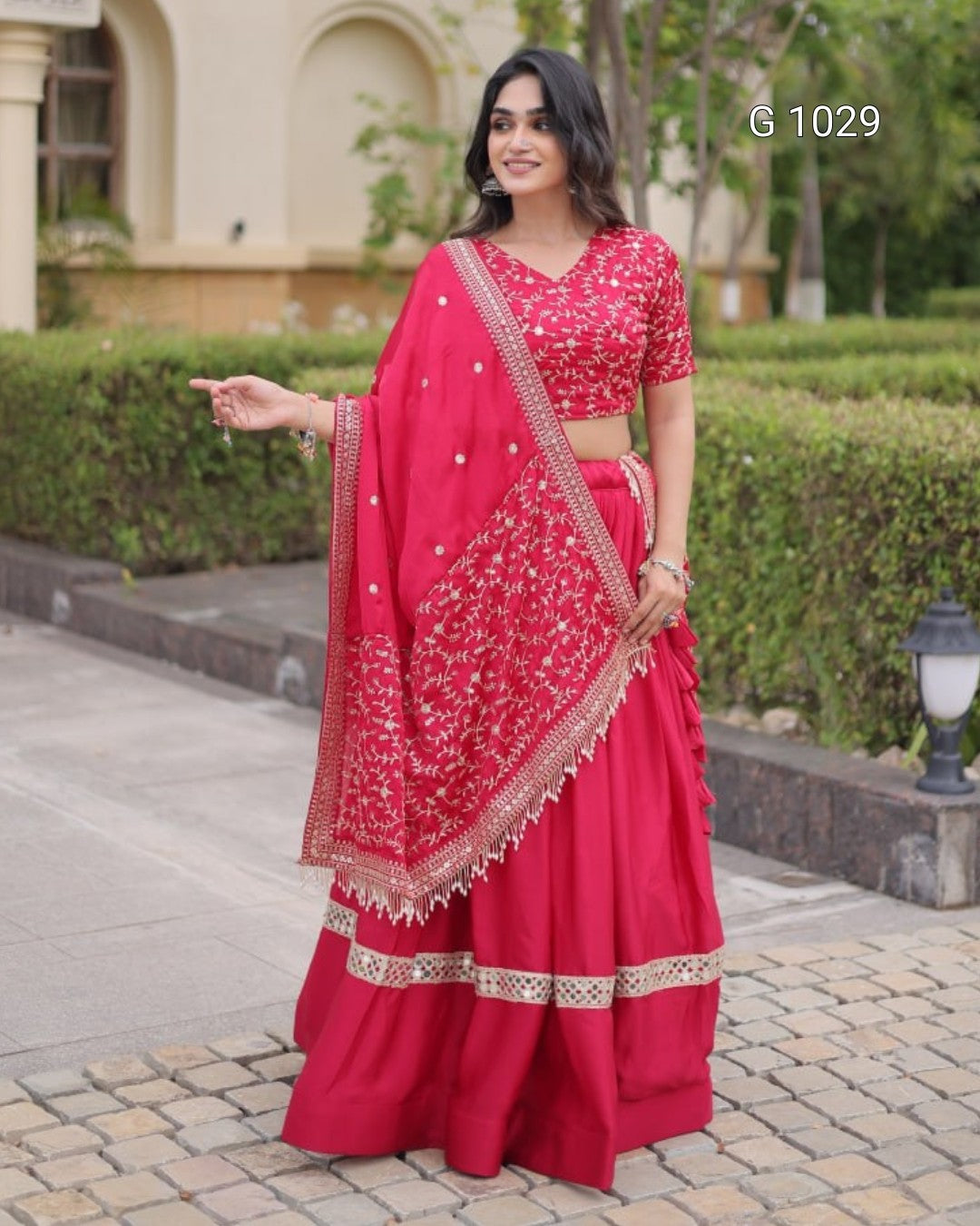 A woman modeling a bright pinkish-red embroidered Indian ethnic outfit, featuring a flared skirt, matching blouse, and draped dupatta with intricate gold and silver mirror work, set against a garden background.