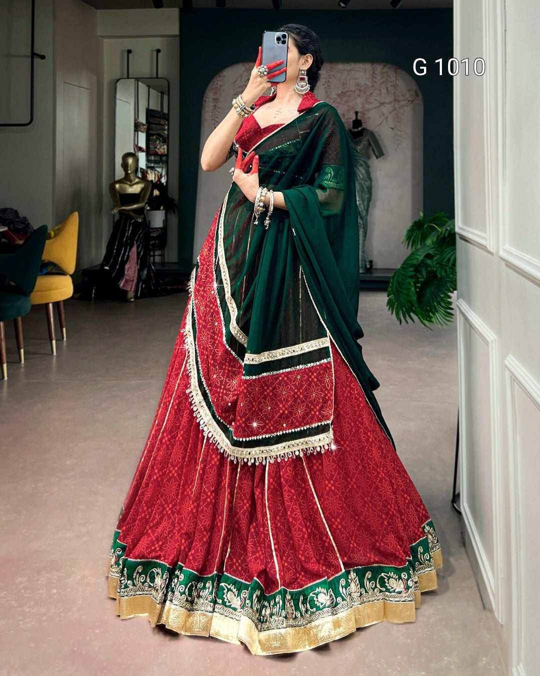 A woman takes a mirror selfie wearing a traditional Indian lehenga choli dress. The dress features a deep red skirt with intricate gold embroidery and a green and black dupatta with gold borders.