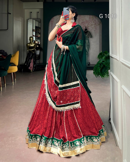 A woman takes a mirror selfie wearing a traditional Indian lehenga choli dress. The dress features a deep red skirt with intricate gold embroidery and a green and black dupatta with gold borders.