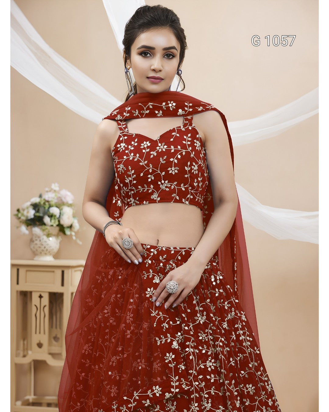 A woman models a red lehenga choli set with white floral embroidery, featuring a sleeveless embroidered choli, matching flared skirt, and draped dupatta, set against a beige background with decorative drapes.
