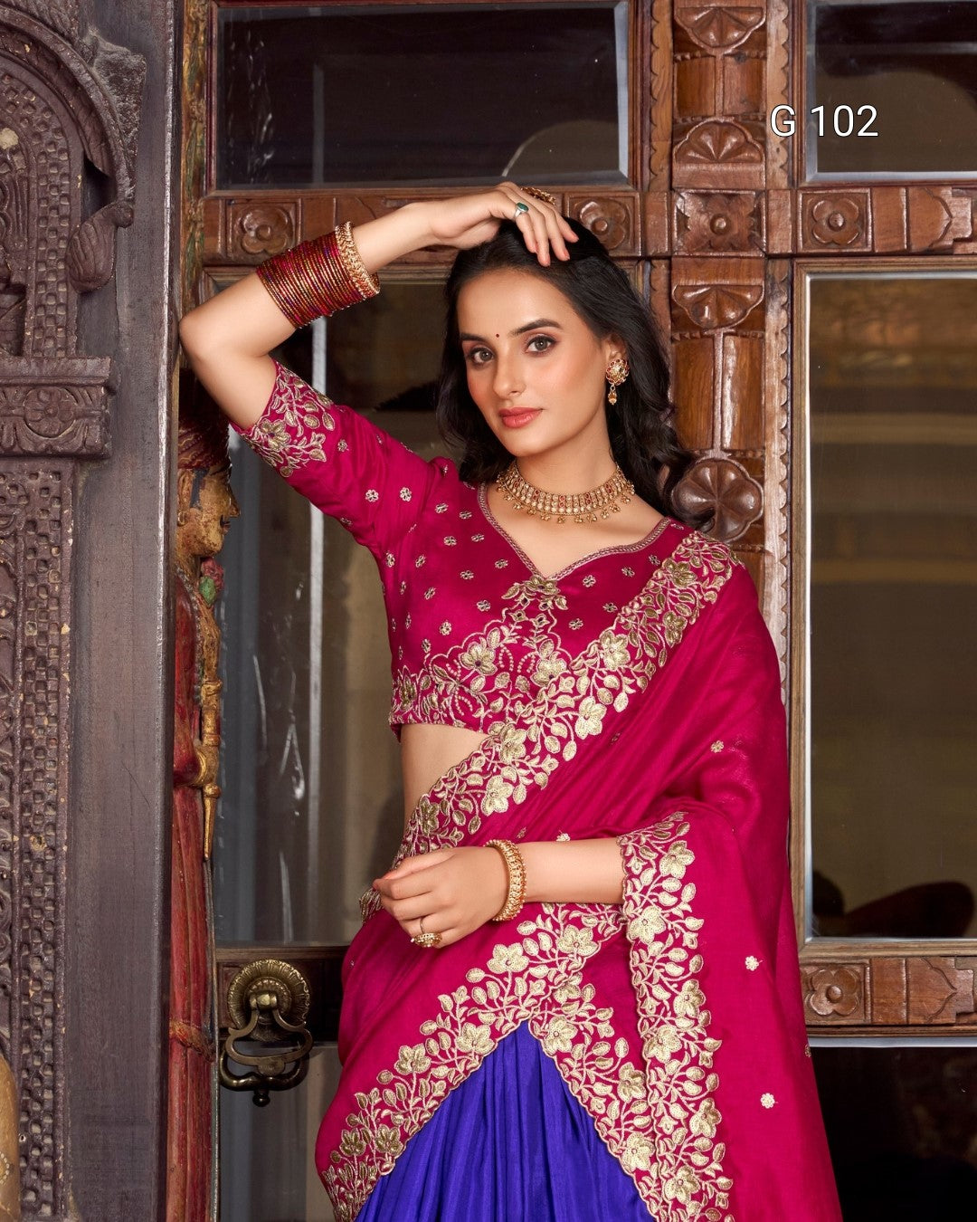 Woman in a traditional pink and blue saree with intricate patterns, standing in front of a wooden door.
