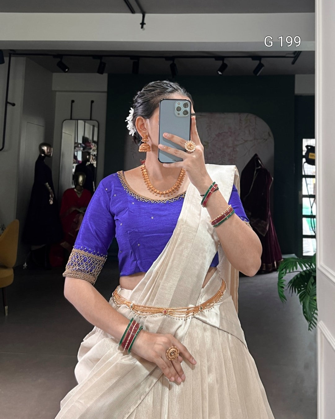 Person wearing a blue blouse and white saree with jewelry, taking a mirror selfie.