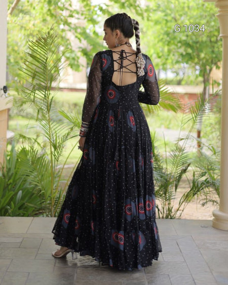 A woman wearing a black long-sleeve maxi dress with red and blue circular motifs and small white dots, shown from the back on a tiled porch with green plants in the background.
