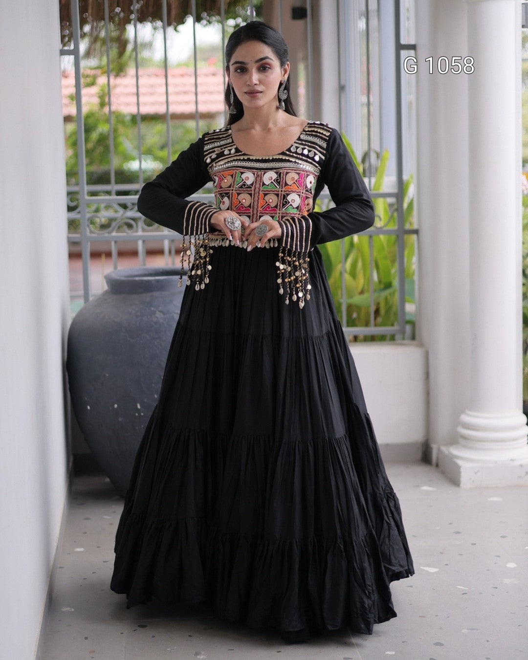 A woman models a black long-sleeve Anarkali gown with a heavily embroidered multicolor neckline and gold-toned embellishments on the sleeves, set against a white-pillared outdoor corridor.