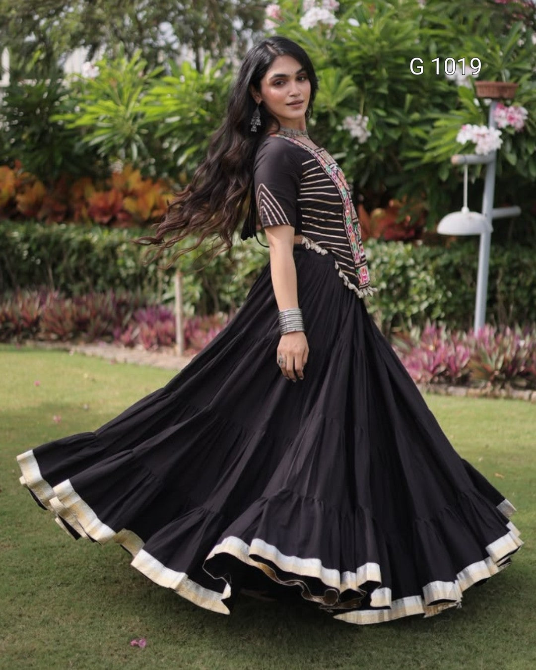 A woman outdoors wearing a black traditional Anarkali-style dress with gold and silver embroidered details on the bodice and a flared skirt with a cream border, captured mid-twirl on a green lawn with garden plants in the background.