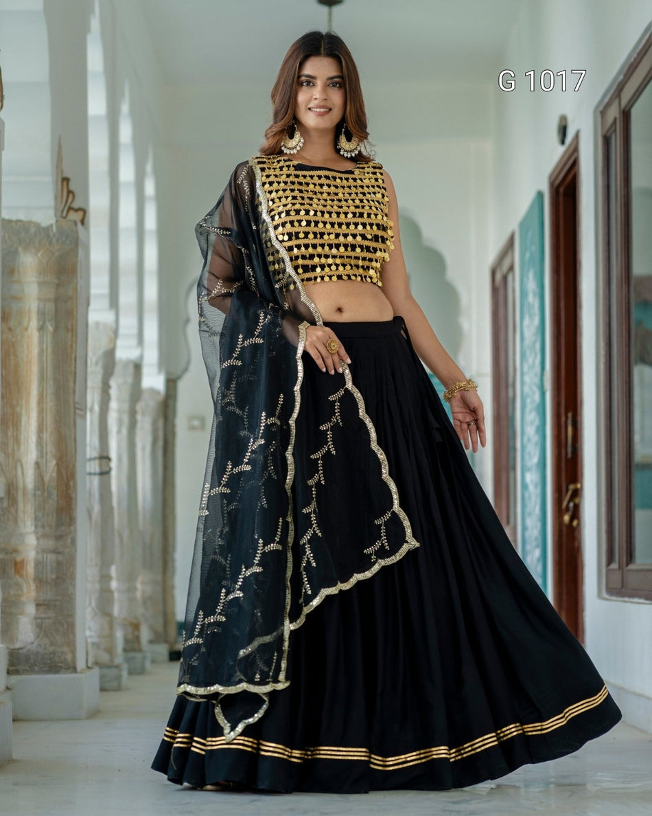 A woman modeling a black and gold traditional Indian lehenga choli set in a corridor. The outfit features a heavily embellished gold crop top, a flowing black skirt, and a sheer black dupatta with gold embroidery.