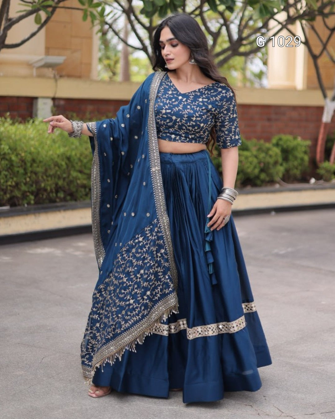 A woman modeling a navy blue traditional Indian lehenga choli set with intricate gold embroidery, standing outdoors on a paved walkway.