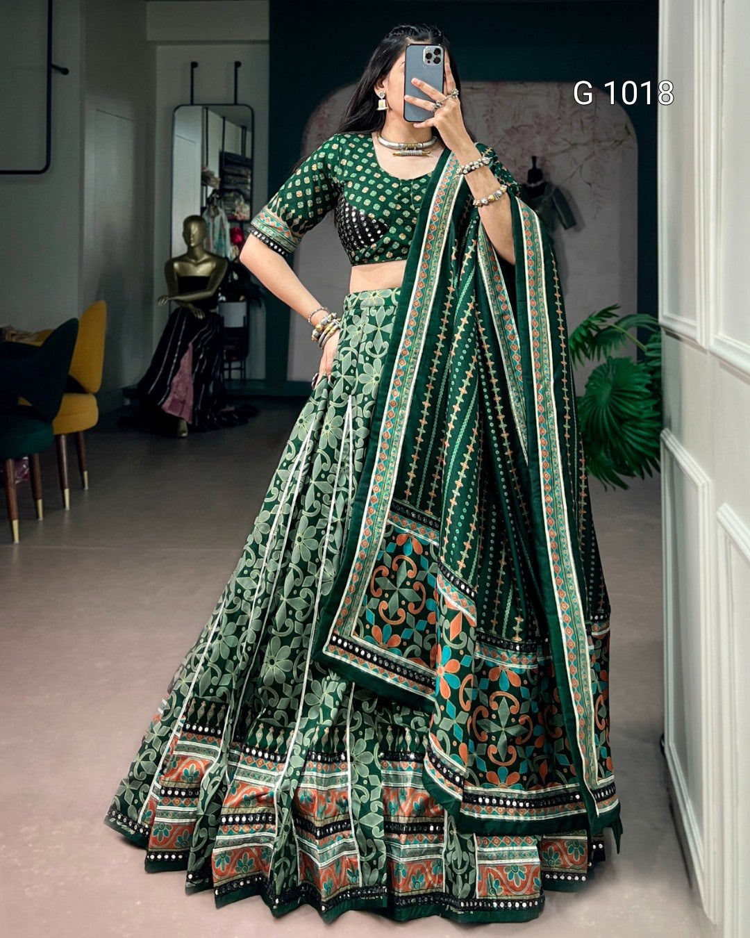 A woman takes a mirror selfie wearing a green and black printed lehenga choli set with a patterned dupatta. The set features intricate floral and geometric designs in green, black, and orange.