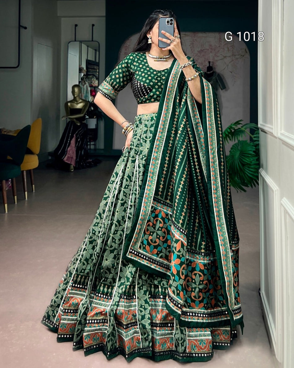 A woman takes a mirror selfie wearing a green and black printed lehenga choli set with a patterned dupatta. The set features intricate floral and geometric designs in green, black, and orange.