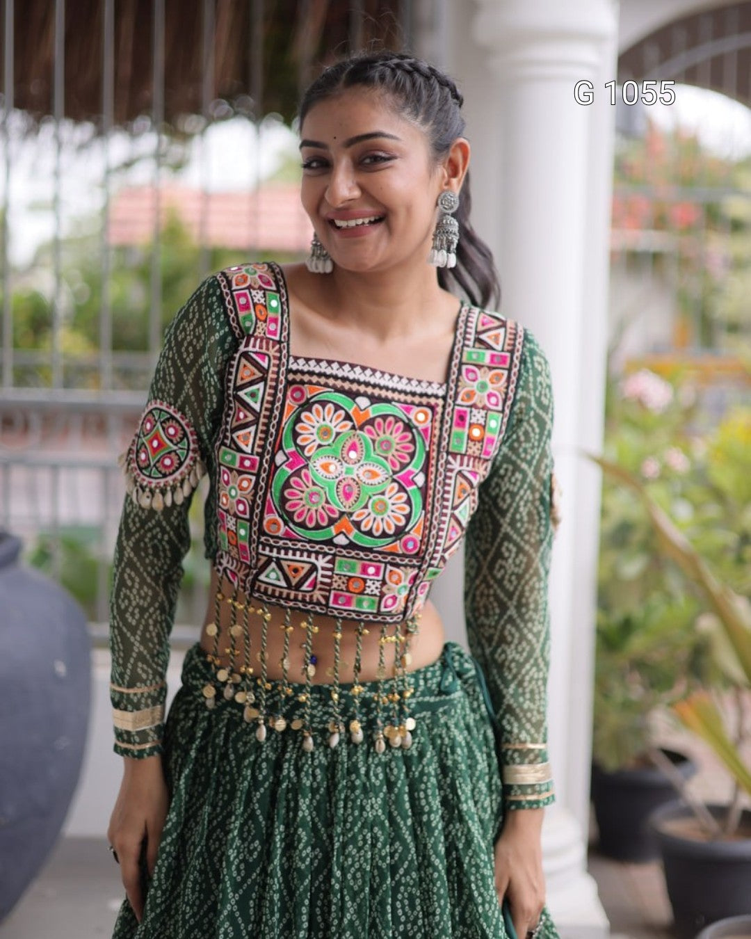 A woman wearing a green embroidered ethnic outfit with a heavily decorated square neckline blouse, matching patterned skirt, and a gold-toned beaded waist chain, standing in front of a white pillar.