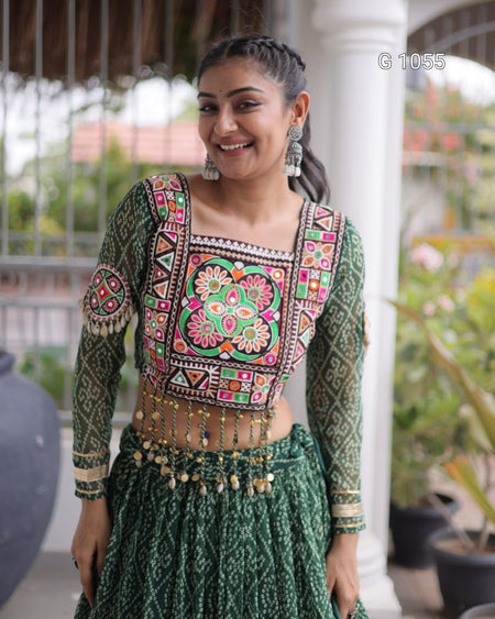 A woman wearing a green embroidered ethnic outfit with a heavily decorated square neckline blouse, matching patterned skirt, and a gold-toned beaded waist chain, standing in front of a white pillar.