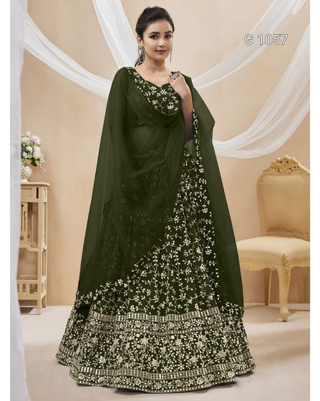 A woman models an olive green ethnic lehenga choli set with intricate white floral embroidery and mirror work, set against a decorated beige background with draped white fabric.