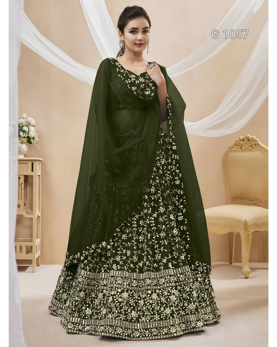 A woman models an olive green ethnic lehenga choli set with intricate white floral embroidery and mirror work, set against a decorated beige background with draped white fabric.