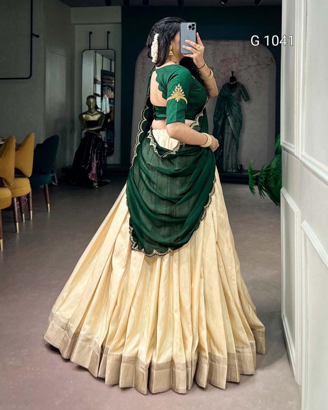 A woman takes a mirror selfie in a boutique wearing a traditional Indian lehenga choli. The outfit features a cream-colored pleated skirt (lehenga) with gold zari borders and a dark green draped blouse and dupatta with gold embroidery.