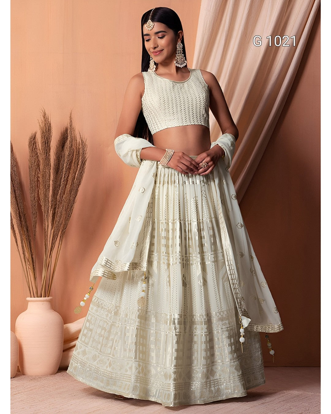 A woman models a traditional Indian outfit consisting of a cream-colored embroidered kurta, matching flared skirt, and sheer sleeves, set against a warm peach-colored backdrop with decorative dried grass in a vase.