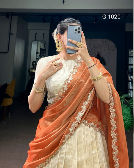 A woman takes a mirror selfie wearing a traditional Indian outfit consisting of a cream-colored textured kurta and a peach-orange dupatta with intricate gold embroidery. She is wearing gold jewelry, including earrings, a ring, and bracelets, and is holding a gray smartphone.