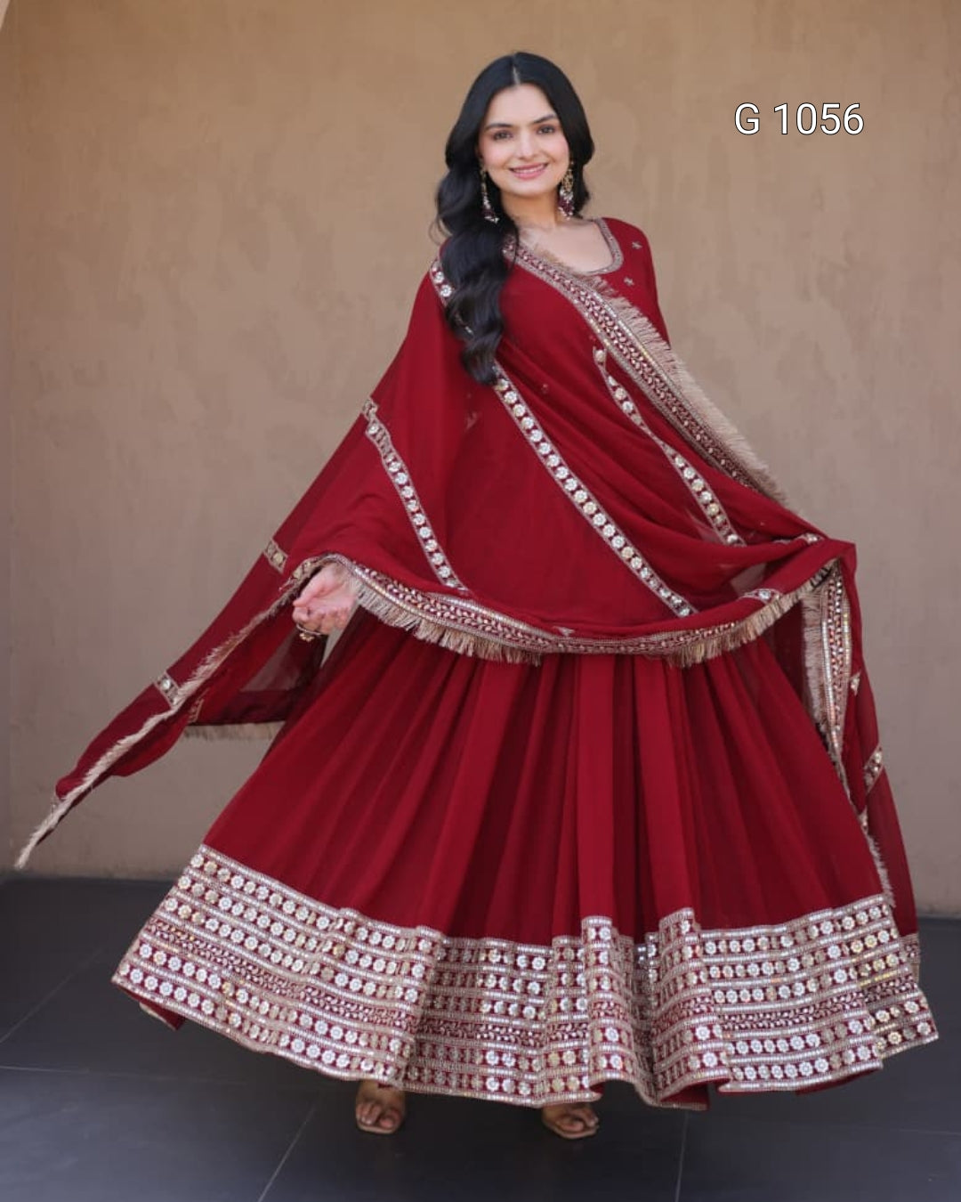 A woman modeling a traditional maroon red sharara suit with intricate gold and silver embroidery, featuring a flared skirt, matching blouse, and dupatta, set against a plain beige wall.