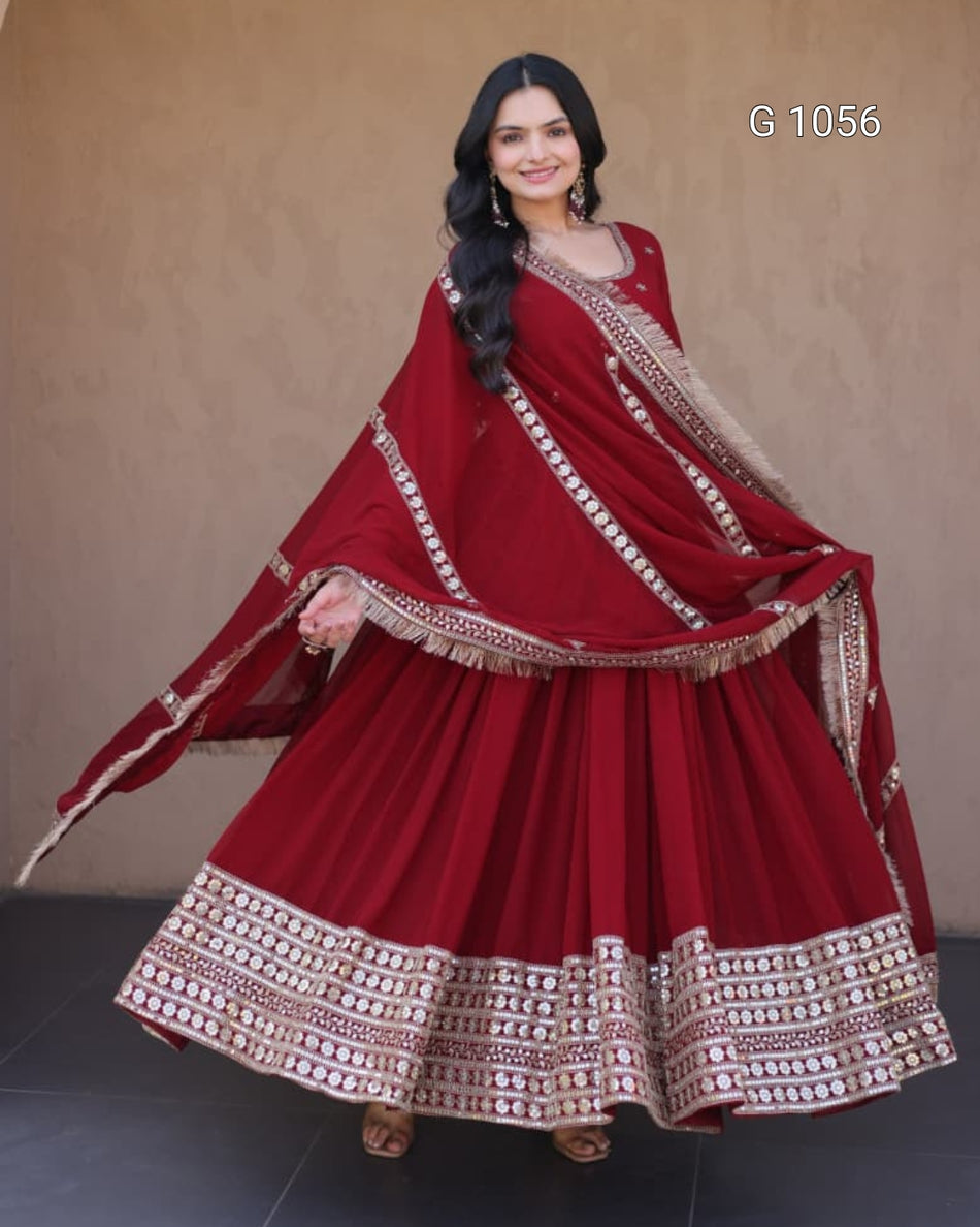A woman modeling a traditional maroon red sharara suit with intricate gold and silver embroidery, featuring a flared skirt, matching blouse, and dupatta, set against a plain beige wall.