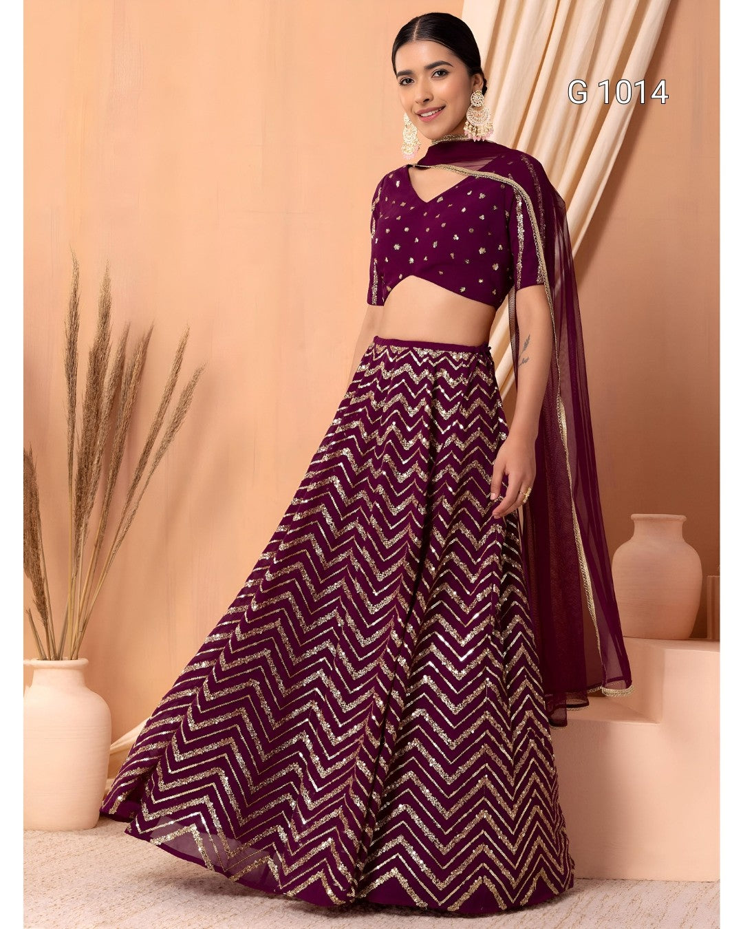 A woman models a deep purple lehenga choli set with gold chevron patterns and a matching sheer dupatta, set against a warm beige background with decorative vases.