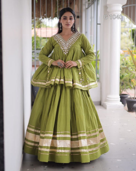 A woman models a traditional Indian anarkali gown in olive green with gold and white embroidery, standing in a corridor with white pillars.