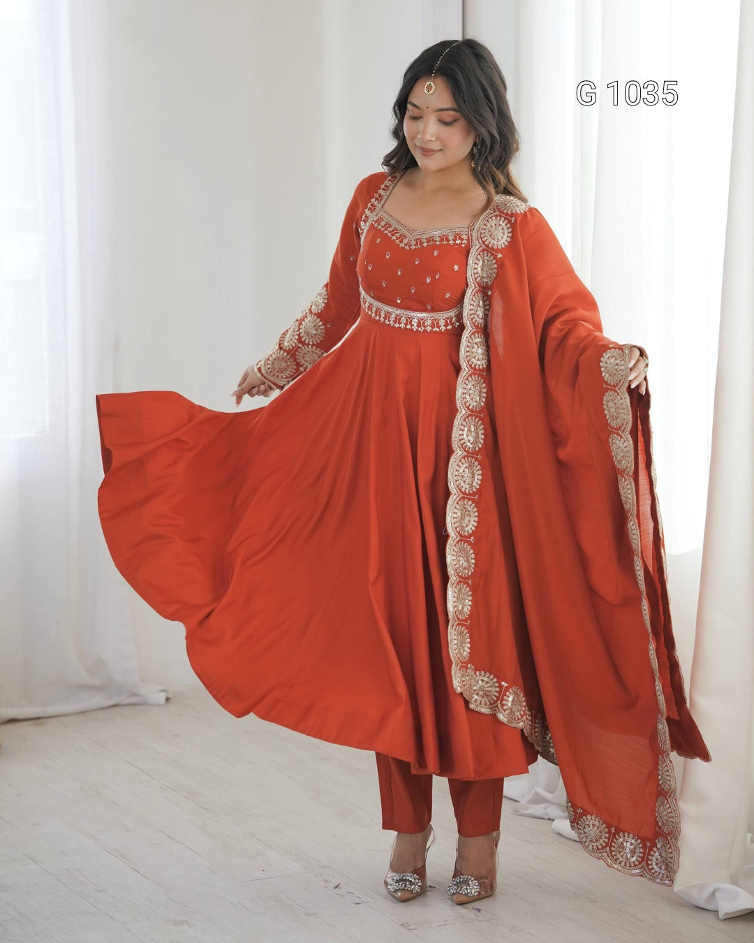 A woman modeling a rust-orange traditional Indian outfit with a matching dupatta, featuring intricate gold embroidery and a flared skirt, standing in a bright room with white curtains.