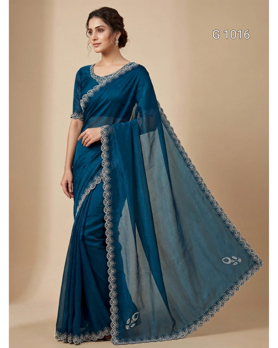 A woman modeling a teal blue saree with a matching blouse, draped elegantly against a solid beige studio background. The saree features a sheer dupatta with a scalloped border and small floral motifs near the lower edge.