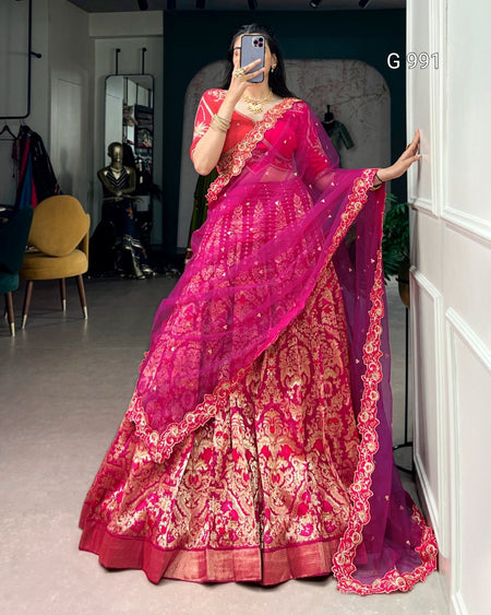 A woman takes a mirror selfie wearing a bright pink and magenta lehenga choli traditional Indian outfit with intricate gold zari embroidery. The setting appears to be a boutique or fitting room with a chair, mirror, and wall visible in the background.