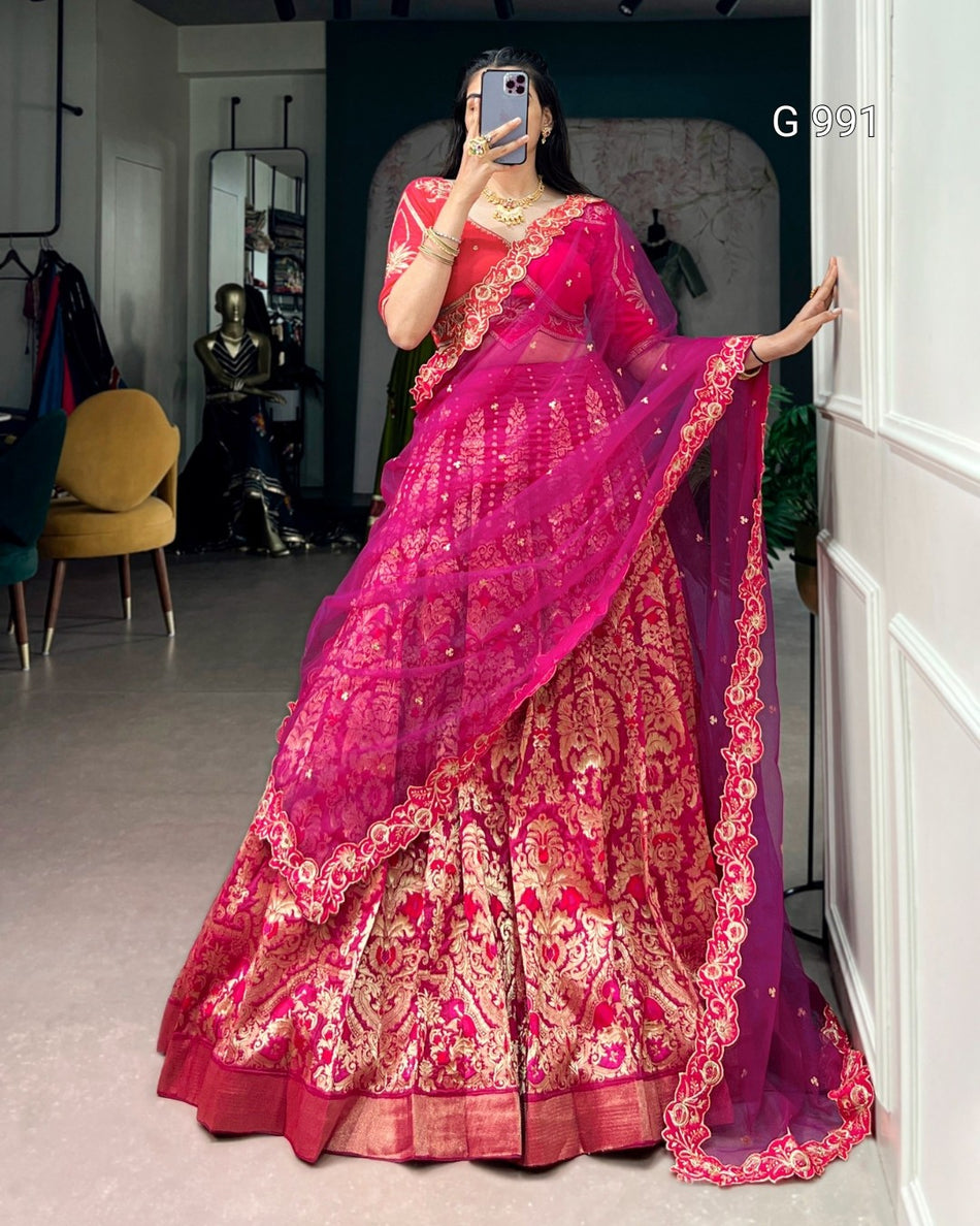 A woman takes a mirror selfie wearing a bright pink and magenta lehenga choli traditional Indian outfit with intricate gold zari embroidery. The setting appears to be a boutique or fitting room with a chair, mirror, and wall visible in the background.