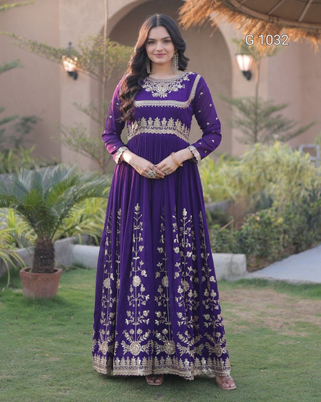 A woman stands outdoors wearing a long purple traditional dress with extensive gold embroidery and embellishments. The dress features a high neckline, long sleeves, and a full skirt with detailed floral patterns.
