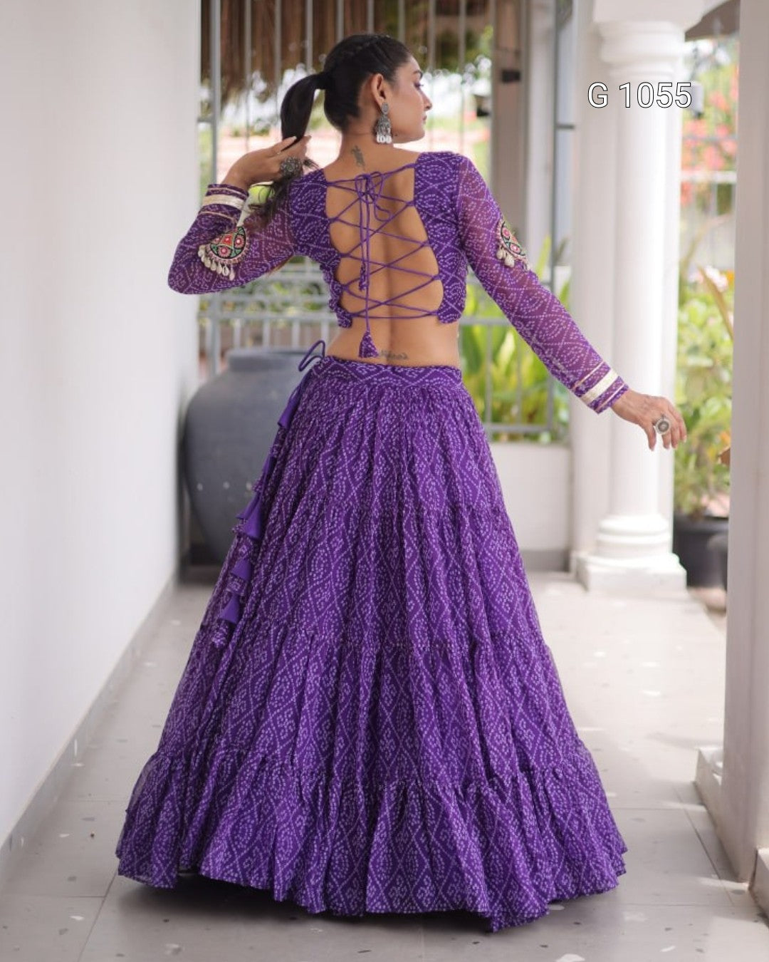 A woman wearing a purple embroidered traditional outfit with a long-sleeve lace-up back blouse and a matching full skirt, standing in a covered walkway.