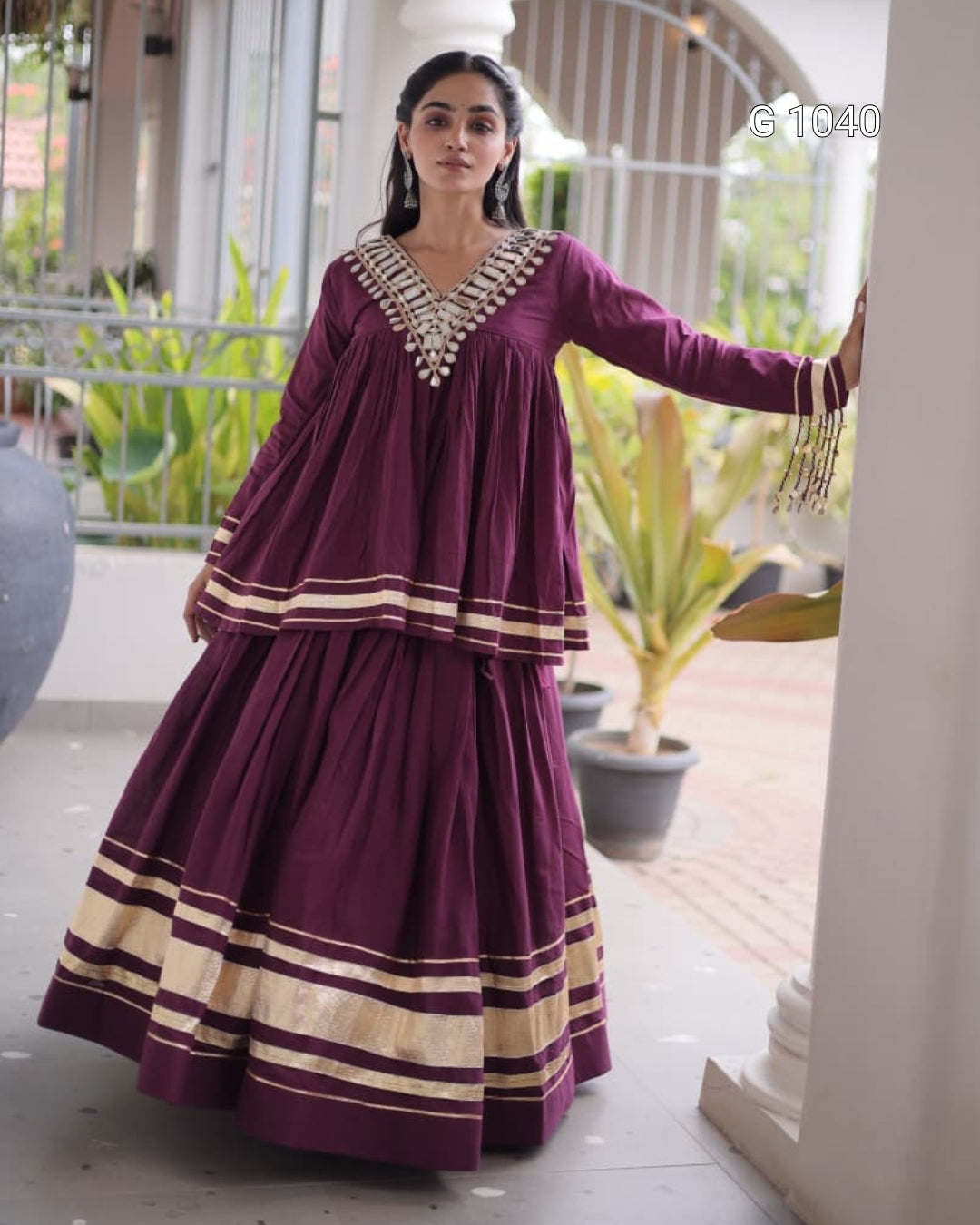 A woman models a deep purple traditional Indian outfit with gold zari stripes and silver embellishments on the neckline, standing in a covered outdoor walkway.
