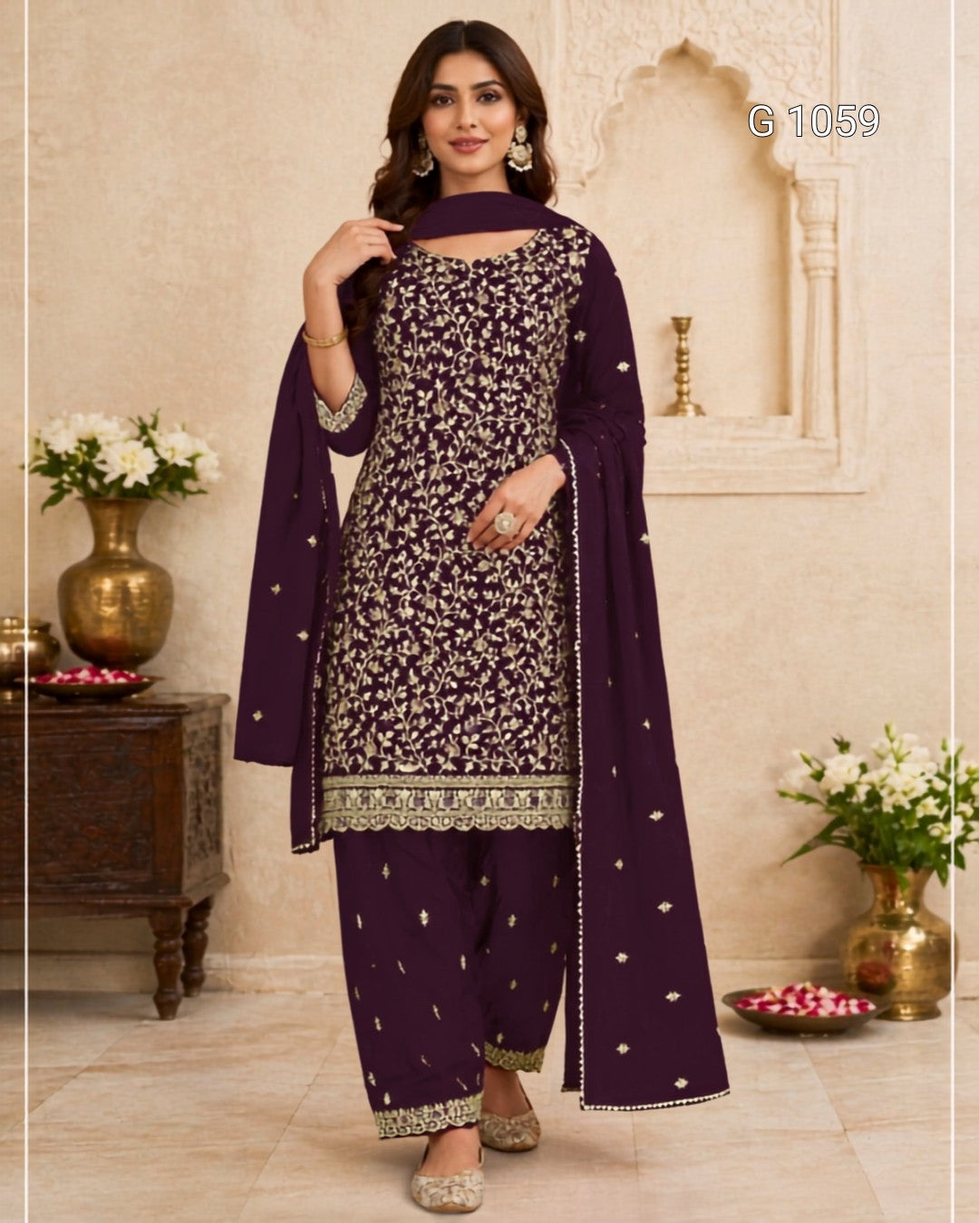 A woman models a deep purple three-piece salwar kameez suit with intricate silver floral embroidery and a matching dupatta, set against a traditional beige architectural background.