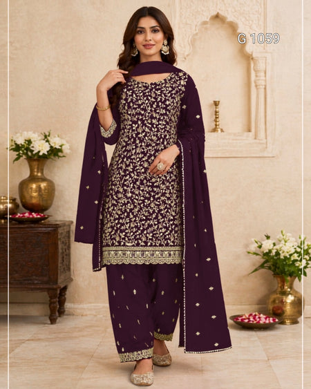 A woman models a deep purple three-piece salwar kameez suit with intricate silver floral embroidery and a matching dupatta, set against a traditional beige architectural background.