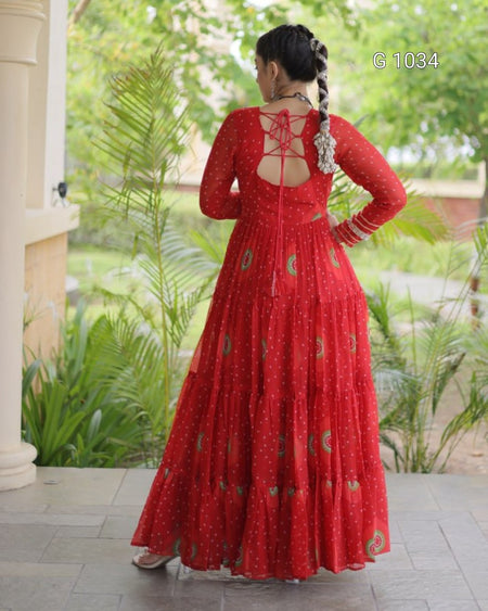 A woman wearing a long red embroidered Anarkali gown, shown from the back in an outdoor garden setting. The dress features a round neckline with criss-cross lace-up back detailing, a flared tiered skirt with small gold and green floral prints, and long sleeves.