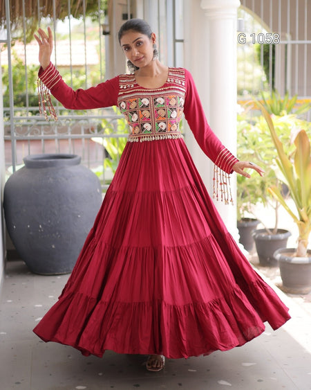 A woman modeling a long-sleeved red Anarkali gown with a heavily embroidered bodice and tiered skirt, standing on a covered patio.