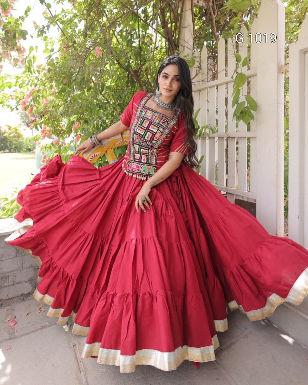 A woman wearing a vibrant red traditional Indian dress with a heavily embroidered neckline and a flared tiered skirt with gold trim, posing outdoors in front of a white fence and green foliage.