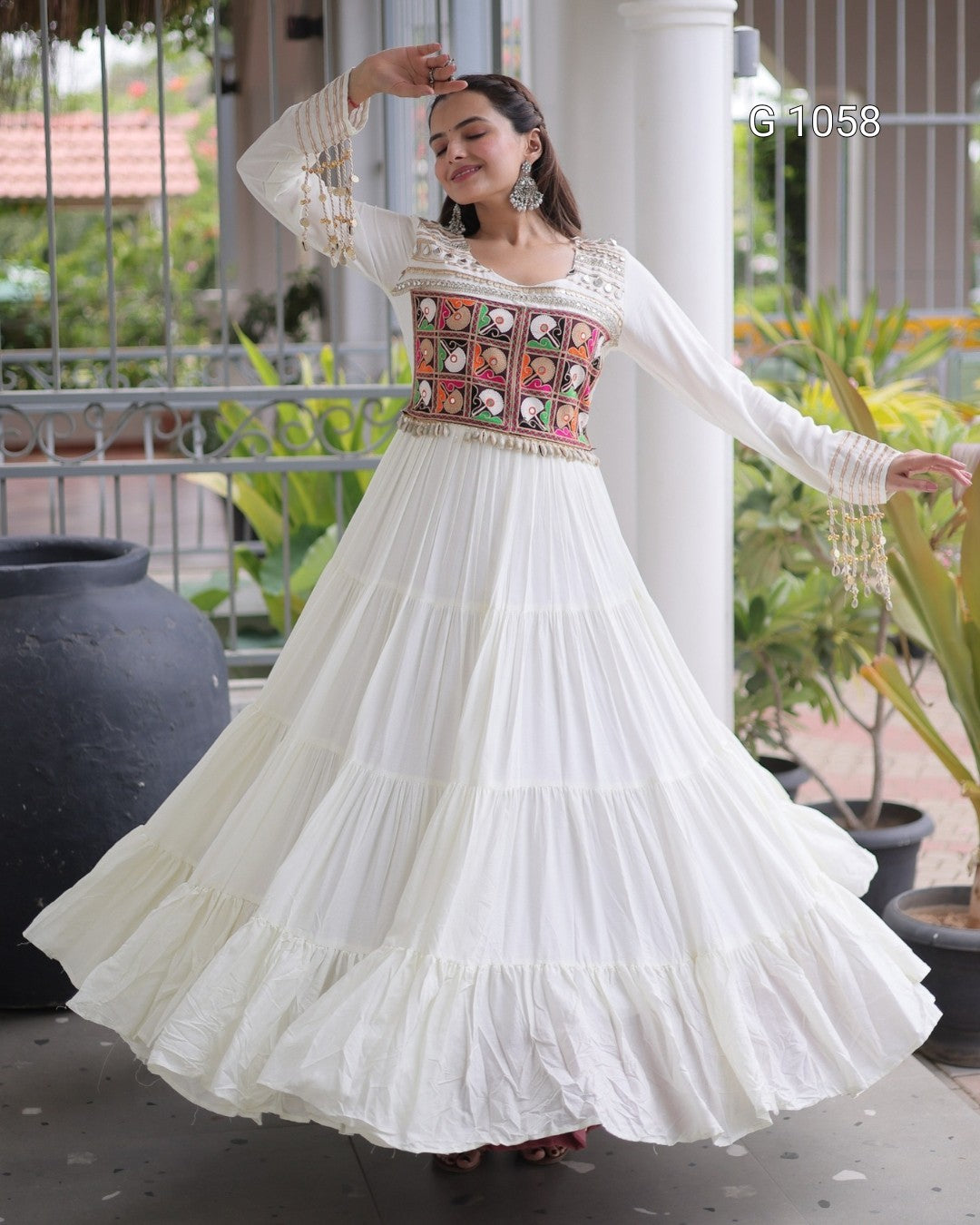 A woman wearing a white tiered anarkali-style dress with a heavily embroidered multicolor bodice, standing on a covered outdoor walkway with plants and columns in the background.