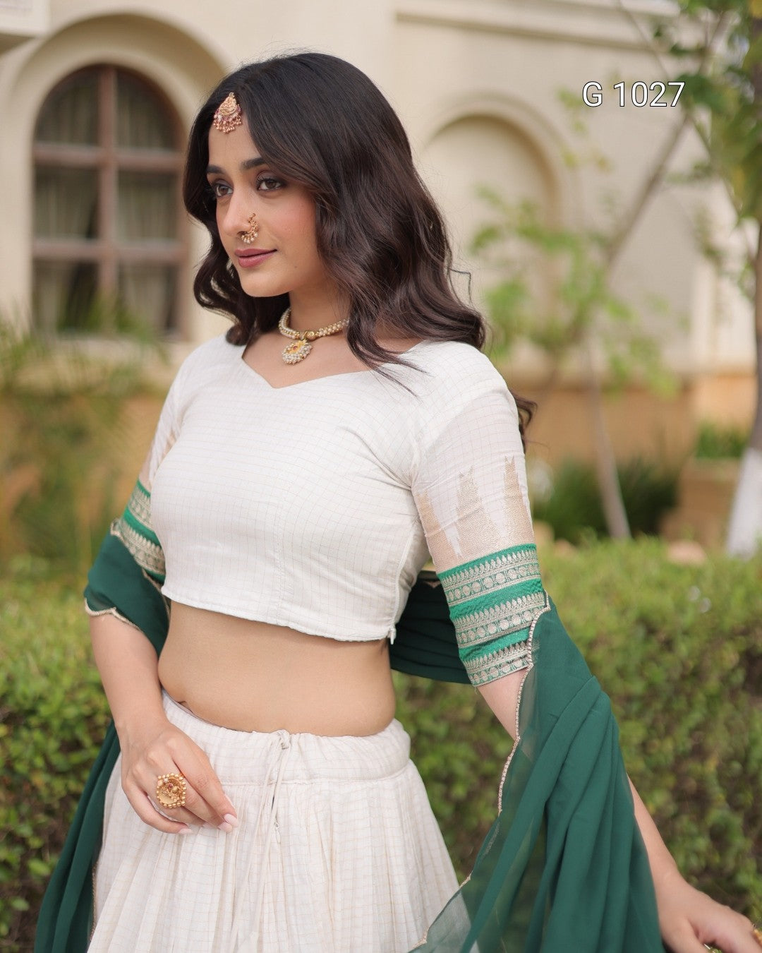A woman wearing a white cropped kurta-style top with sheer sleeves and green patterned trim, paired with a white skirt and a dark green dupatta, posed outdoors in front of a blurred building and garden background.